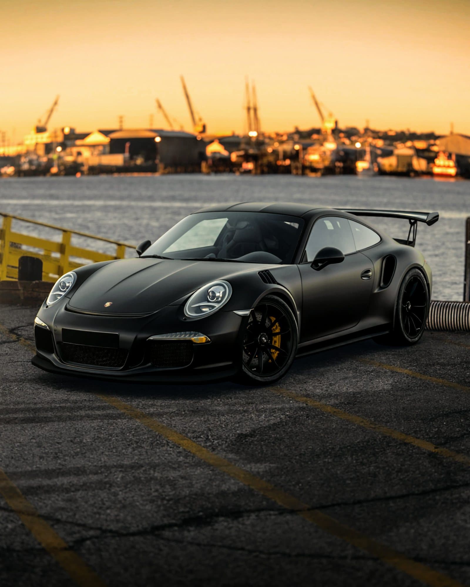 Matte black Porsche 911 with a large rear wing at a dockside at dusk